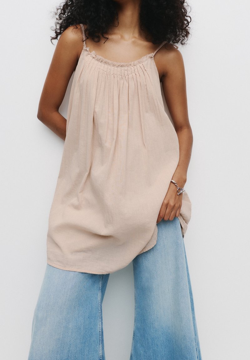 Femme portant un top beige ample sans manches et un jean bleu clair à jambes larges, posant devant un fond blanc uni.