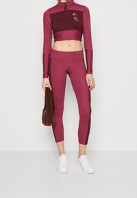 Burgundy träningsset som innehåller en cropped långärmad topp med dragkedja och matchande leggings. Vita skor fulländar looken.