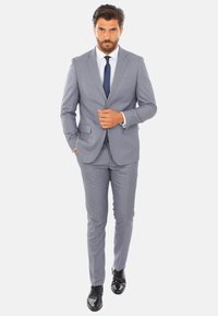 Completo grigio chiaro con vestibilità slim, rever a scollo a V e chiusura con un solo bottone. Abbinato a una camicia bianca e una cravatta scura. Scarpe nere lucide.
