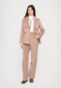 Braune Cord-Jacke mit doppelter Knopfleiste und passenden Hosen, kombiniert mit einem weißen Hochkragenoberteil und einer Bolo-Krawatte. Das Modell steht vor einem schlichten Hintergrund.