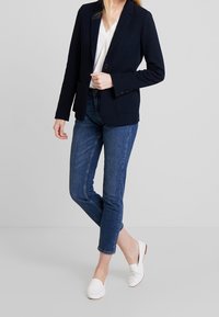 Granatowa marynarka w prążki z teksturowanej tkaniny, zestawiona z ciemnymi dżinsami. Białe płaskie buty dopełniają stylizację. Tło jest neutralne.