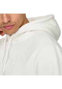Sweatshirt blanc avec une capuche à cordon, texture lisse, coutures renforcées et détails de couture visibles sur les épaules et les manches.