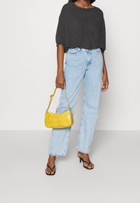 Schwarzes, übergroßes, kurz geschnittenes Oberteil; hellblaue, gerade geschnittene Jeans; gelbe Handtasche aus lackiertem Leder mit kurzem Tragegurt; schwarze, Riemchen-Sandaletten.