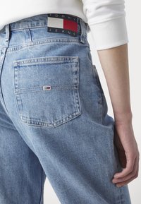 Primer plano de una persona que lleva unos vaqueros Tommy Jeans de color azul claro con un parche de logo en la cinturilla y una pequeña bandera bordada en el bolsillo trasero.