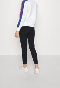 Vitt sweatshirt med röda och blå ränder på axlarna, tillsammans med svarta leggings och vita sneakers, visad bakifrån.