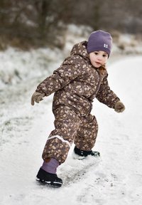 Combinaison de neige marron à motifs floraux avec une fermeture éclair à l'avant, une bande réfléchissante blanche et un chapeau violet ; portée avec des moufles et des bottes noires sur la neige.