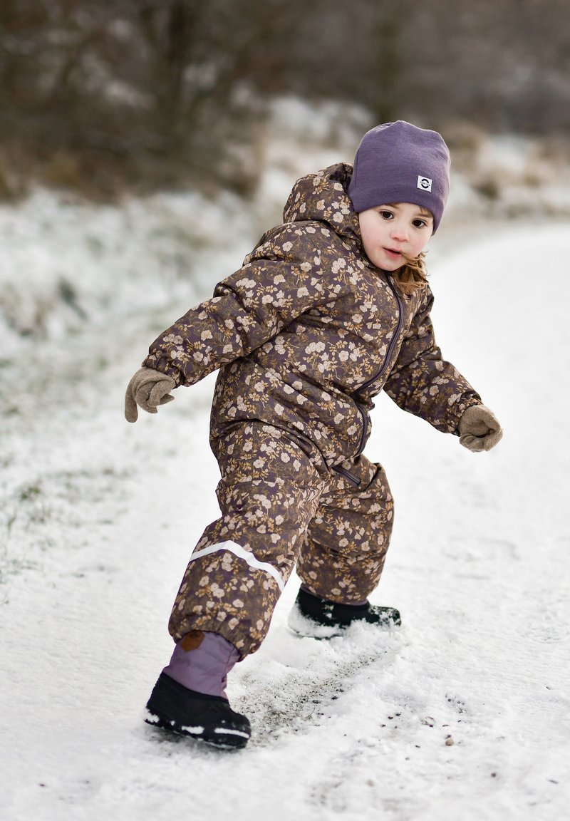 Combinaison de neige marron à motifs floraux avec une fermeture éclair à l'avant, une bande réfléchissante blanche et un chapeau violet ; portée avec des moufles et des bottes noires sur la neige.