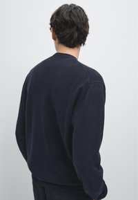 Námořnický fleece svetr se kulatým výstřihem, žebrovanými manžetami a uvolněným střihem. Textura se zdá být měkká s jemným zrnitým povrchem.