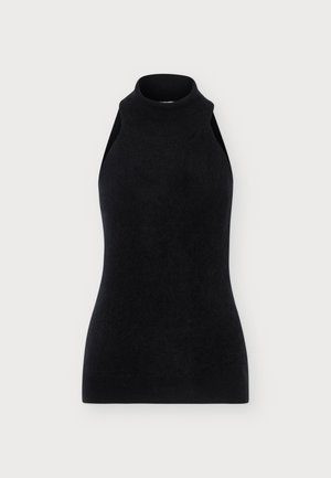 Femme portant un haut noir sans manches à col roulé, une longue jupe vert olive et des bottes noires à talons, debout devant un fond clair uni.