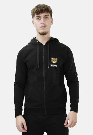 Felpa con zip nera realizzata in tessuto morbido, con un logo di un orso e un branding a contrasto sul lato sinistro del petto. Include un cappuccio e tasche frontali.
