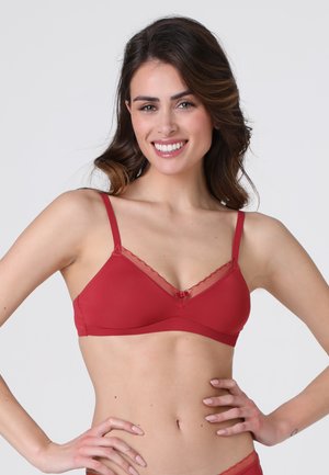 Donna sorridente con capelli lunghi che indossa un reggiseno rosso con spalline sottili regolabili e bordo trasparente, con le mani posate sui fianchi.