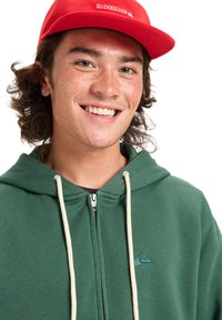 Groene rits hoodie met crèmekleurige koorden en een klein blauw logo. Rode pet met het "QUIKSILVER" logo. Jongeman met krullend haar en een glimlach.