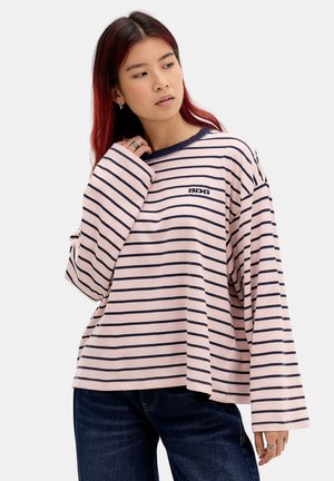 Jeune femme aux cheveux roux portant une chemise ample à rayures roses et bleu marine à manches longues et un jean foncé sur un fond uni.