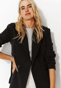 Blazer noir ajusté avec deux boutons à l'avant, revers crantés et manches retroussées, superposé sur un t-shirt gris. Texture lisse.