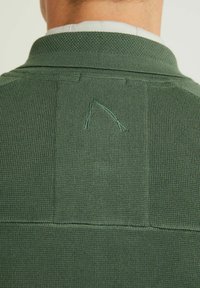 Grön stickad polo t-shirt med ett strukturerat mönster och en liten synlig tråd vid bakhalsen. Tyget verkar mjukt och andningsbart.