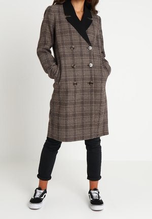 Kobieta ubrana w sięgający do kolan brązowy płaszcz w kratę z podwójnym rzędem guzików i czarnymi klapami, czarne podwinięte spodnie oraz czarno-białe tenisówki.
