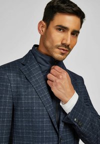 Marineblauer karierter Blazer mit einem Reißverschluss und hohem Kragen als Innenschicht. Glattes Material, kontrastierender weißer Manschettenbereich, Fünf-Knopf-Detail an den Ärmeln.