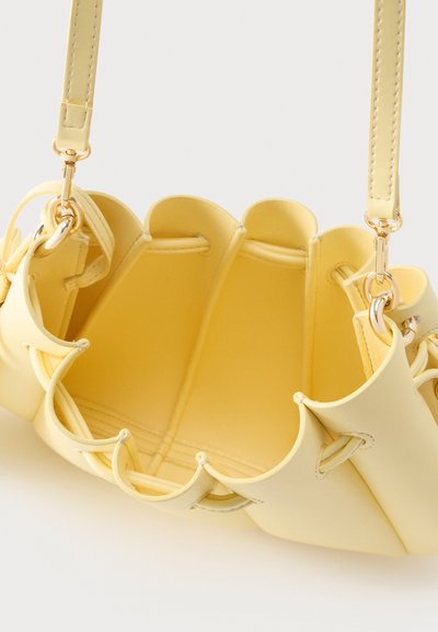 Μικρή κίτρινη δερμάτινη τσάντα ώμου με κυματιστές άκρες, ανοιχτή κορυφή, ραμμένα πάνελ και μεταλλικά στοιχεία σε χρυσό τόνο.