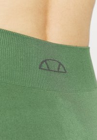 Close-up of a person’s lower back wearing green ribbed fabric leggings with a small black semi-circle logo on the waistband.
