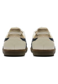 Bílé kožené tenisky s béžovými akcenty a námořnickým bočním pruhem, vybavené texturovanou gumovou podrážkou a zaobleným designem zadní části.