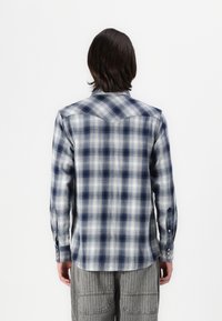 Camicia in flanella a quadri blu e bianca con maniche lunghe, polsini con bottoni e orlo curvo; dettaglio della yoke visibile sulla schiena.