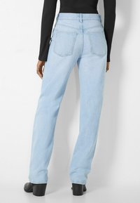 Person in hellblauen, geraden Jeans, schwarzen High-Heels und einem langen schwarzen Rippoberteil, das vor einem weißen Hintergrund steht.