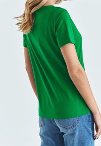 T-shirt di cotone verde con maniche corte, scollo rotondo e vestibilità comoda. La vista posteriore mostra una texture liscia e un design semplice. Visibili anche jeans in denim.