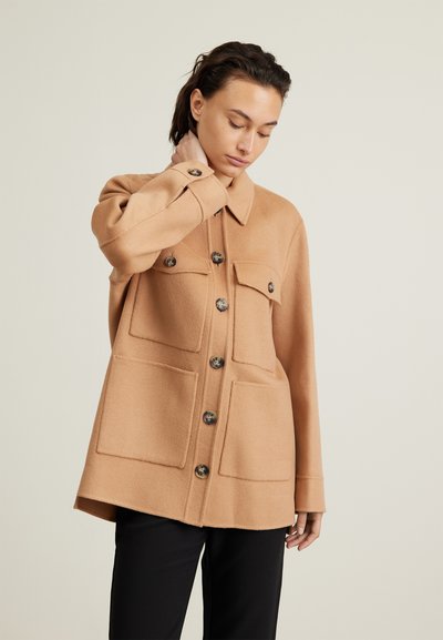 Frau, die sich am Hals berührt, trägt eine camelartige Knopfjacket mit vier großen Taschen und schwarze Hosen vor einem schlichten hellen Hintergrund.