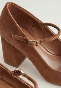 Brown suede high-heeled shoes with a square heel, featuring a buckle strap across the instep and a smooth inner lining.