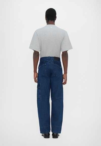 Camiseta gris de manga corta y pantalones vaqueros oscuros de pierna ancha con múltiples bolsillos y una cinturilla negra, vistos desde la parte de atrás sobre un fondo blanco.