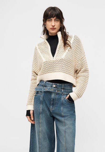 Γυναίκα με σκούρα μαλλιά φοράει κρεμ ανοιχτό πλεκτό cropped πουλόβερ πάνω από μαύρο τοπ και τζιν μπλε με ψηλή μέση και φαρδιά μπατζάκια με διπλή ζώνη μέσης.