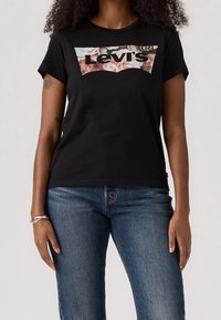 Camiseta negra de Levi's con logotipo floral, llevada por una persona con cabello rizado, jeans azules y una pulsera plateada en la muñeca izquierda.