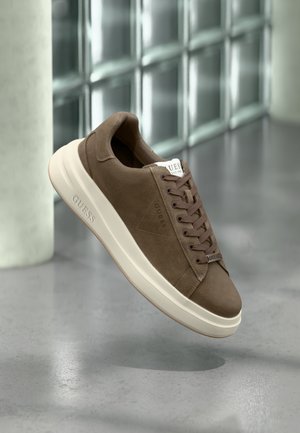 Brown suede Guess sneaker with white sole, floating above a gray polished floor beside glass block wall.