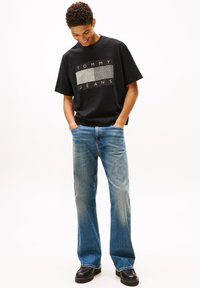 Camiseta negra de manga corta con un logo rectangular gris texturizado que dice "TOMMY JEANS", combinada con jeans de pierna recta en azul claro y zapatos negros.