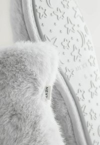 Harmaat pehmeät tohvelit, joissa on pehmeä pinta ja valkoinen pohja, johon on painettu tähti- ja kuukuvioita, ja sivussa näkyy merkkilappu.