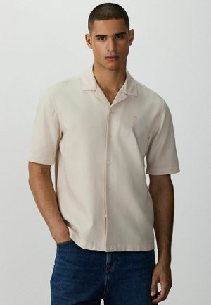 Uomo con capelli corti che indossa una camicia beige a maniche corte con bottoni e jeans blu, in piedi con una mano in tasca contro uno sfondo semplice.