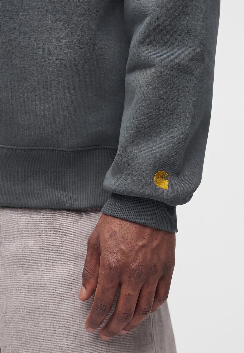 Dunkelhäutige Hand ruht seitlich, trägt dunkelgrauen Sweatshirt mit gelbem Carhartt-Logo am Ärmelsaum und hellgraue Hose.