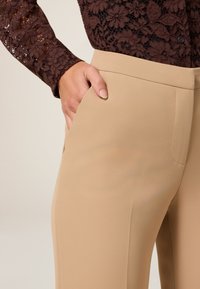 Beige op maat gemaakte broek met een gladde stof, uitgerust met zijzakken en een rechte pijpdesign. Draagt men met een donker kant blousje.