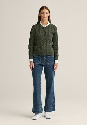 Kvinde stående iført en grøn kabelstrikket knaptrøje, hvid skjorte, blå vidde jeans og hvide sneakers mod en ensfarvet baggrund.