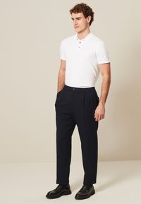 Camisa polo blanca con tres botones, combinada con pantalones oscuros de pinzas color navy y zapatos negros, presentada sobre un fondo neutral.