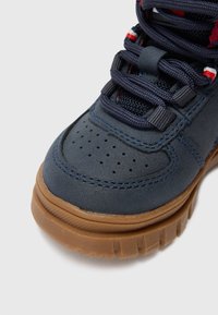 Zapatilla azul marino con un upper texturizado, puntera perforada, diseño de cordones y suela de goma marrón. Presenta acentos rojos y blancos en los cordones.