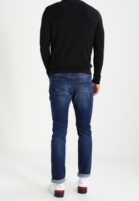 Dunkelbl sweater, blaue Jeans mit heller Umschlagkante. Weiße Sneakers mit roten Akzenten. Ansicht von hinten, die Passform und Stilmerkmale zeigt.