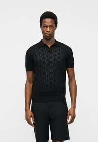 Άνδρας που φοράει μαύρο πόλο με κοντά μανίκια και διακριτικά υφασματικά μοτίβα, καθώς και μαύρη βερμούδα, στέκεται μπροστά σε απλό λευκό φόντο.