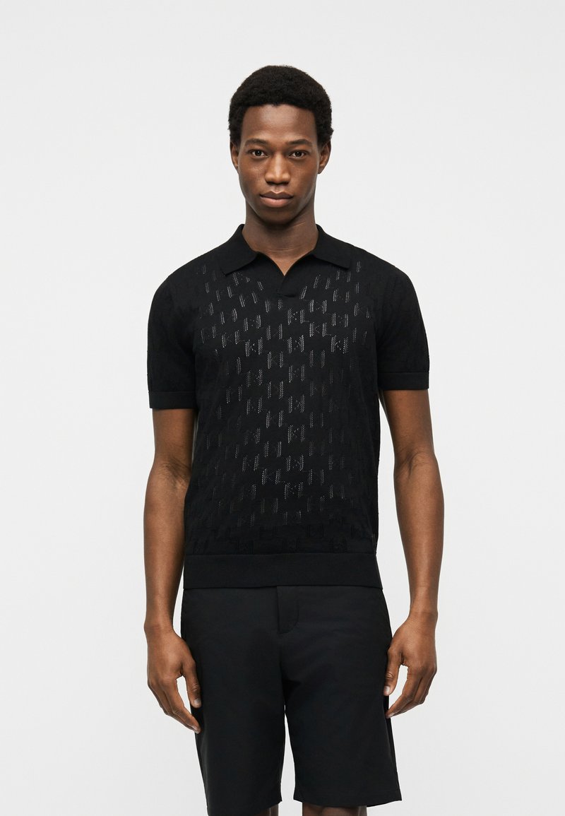 Άνδρας που φοράει μαύρο πόλο με κοντά μανίκια και διακριτικά υφασματικά μοτίβα, καθώς και μαύρη βερμούδα, στέκεται μπροστά σε απλό λευκό φόντο.