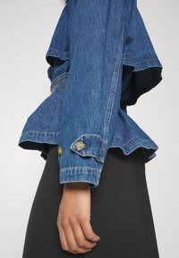 Jeansjacka med en utsvängd design, blå färg och uppvikta ärmar. Har knappar som detalj och en volangnederkant. Materialet verkar texturerat.