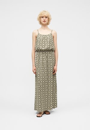 Person iført ærmeløs, mønstret maxikjole med geometrisk blomsterdesign og sorte sandaler, stående foran en ensfarvet lys baggrund.