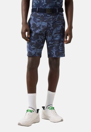 Mann trägt marineblaue Shorts mit Camouflage-Muster und Palmen, einen schwarzen Gürtel, weiße Socken und weiße Turnschuhe mit grünen Schnürsenkeln.