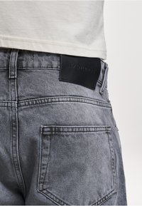 Gray denim jeans with a textured finish, featuring a black leather patch with embossed branding at the back and standard pocket design.
