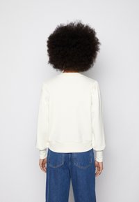 Felpa bianca con scollo rotondo, polsini e orlo a coste. Dettagli plissettati sulle spalle, design leggermente cropped, abbinata a jeans blu.
