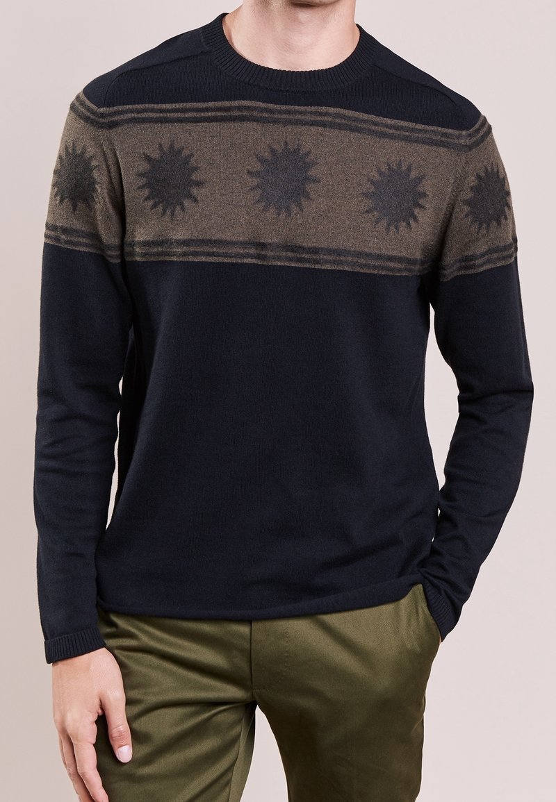 Homme portant un pull à manches longues noir et olive avec une bande de motifs rayons de soleil sur la poitrine, associé à un pantalon olive.